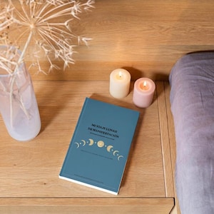 Op de afbeelding: Een blauw boek met de titel MI VIAJE LUNAR DE MANIFESTACIÓN met een gouden maanfase-afbeelding op de kaft, geplaatst naast een doorzichtige glazen vaas met een gedroogde alliumbloem en twee kleine brandende kaarsen, een witte en een lichtroze.
