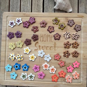 May include: A collection of small, five-petaled flower-shaped buttons in various colours and patterns, including polka dots, leopard print, stripes, and solid hues. The buttons are arranged on a wooden board with the text Home Sweet Home engraved.