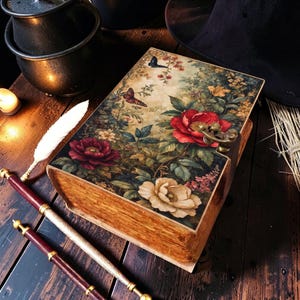 Pode incluir: Uma caixa retangular de madeira projetada para se assemelhar a um livro antigo, com uma estampa floral na tampa apresentando flores vermelhas, brancas e amarelas, folhagem verde e borboletas. A caixa inclui um fecho de metal cor de latão.