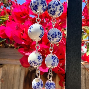 May include: A pair of dangle earrings featuring four stacked white ceramic beads on each side, decorated with a blue floral pattern. The beads are connected by silver-toned metal jump rings and capped with small silver-toned metal flower-shaped findings.