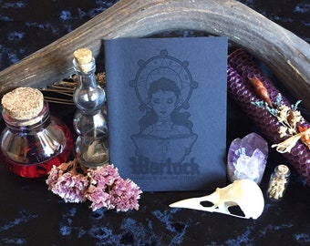 Puede incluir: Un diario negro con una ilustración en relieve plateado de una mujer leyendo un libro. El diario tiene el texto "Warlock" y "WITCHY PLANNER COVER" en la portada.