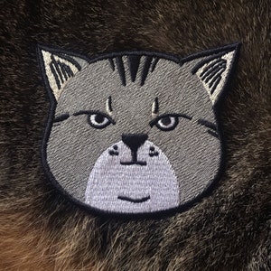 May include: Embroidered gray cat patch with black eyes and a white nose and mouth. The cat has a serious expression.