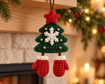 Op de afbeelding: Een gehaakte kerstboomhanger bestaande uit een donkergroene driehoekige boom met een rode ster aan de top, een witte sneeuwvlok in het midden en kleine gouden kralen. Twee rode gehaakte wanten met witte boorden hangen onderaan de boom.