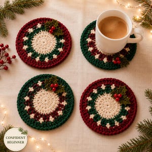 May include: Four round crochet coasters featuring a cream centre, a dark green and maroon border, and a small green holly leaf applique with three red berries. One coaster supports a white ceramic mug filled with a light brown beverage.