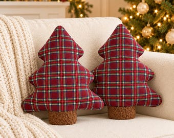 Op de afbeelding: Twee decoratieve kussens in de vorm van kerstbomen, voorzien van een rood, groen en wit ruitpatroon. Elk kussen heeft een gestructureerde, bruine, pluizige basis die een boomstam voorstelt, wat zorgt voor een zachte uitstraling.
