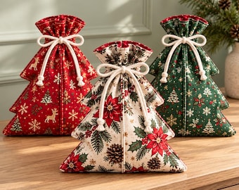 Op de afbeelding: Drie boomvormige stoffen cadeauzakjes met trekkoorden. Het linkerzakje is rood met gouden rendieren en sneeuwvlokken. Het middelste zakje is crème met rode kerststerren en dennenappels. Het rechterzakje is groen met witte sneeuwvlokken.