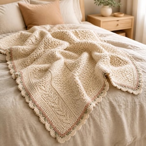 May include: A cream-colored crocheted blanket featuring a textured stitch pattern with a central leaf-like design. The edges are finished with a scalloped border in cream, accented by thin lines of soft pink and sage green yarn throughout the trim.