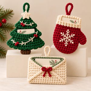 Puede incluir: Tres adornos de ganchillo que incluyen un árbol de Navidad verde oscuro con una estrella blanca, un guante rojo con un copo de nieve blanco y un sobre color crema con un lazo rojo. Cada pieza tiene un lazo para colgar y una tarjeta.