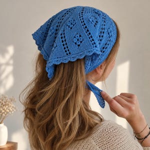 May include: A bright blue triangular headscarf crafted with a detailed knit pattern featuring small floral motifs and openwork sections. The headscarf has a scalloped edge and includes thin knitted ties at the corners to secure it in place.
