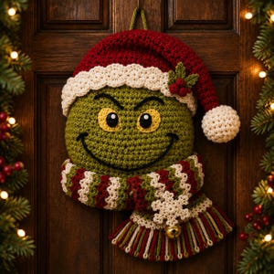 May include: A round, green, crocheted character head with yellow eyes and a black stitched smile. It wears a red and white crocheted Santa hat with a holly accent and a striped red, white, and green scarf featuring a white snowflake and a gold bell.