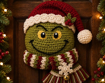 Op de afbeelding: Een rond, groen, gehaakt karakterhoofd met gele ogen en een zwarte geborduurde glimlach. Het draagt een rood-witte gehaakte kerstmuts met een hulstaccent en een gestreepte rood-wit-groene sjaal met een witte sneeuwvlok en een gouden belletje.