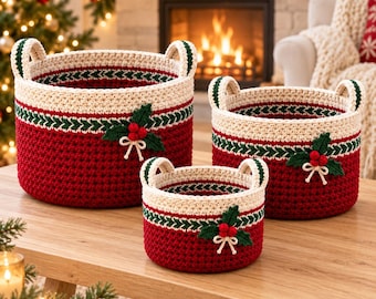 Op de afbeelding: Een set van drie ronde, gehaakte opbergmanden in verschillende maten. Elke mand heeft een dieprode basis, een crèmekleurig bovengedeelte en een groene decoratieve band. Een gehaakte hulstapplicatie met rode bessen siert de voorzijde van elke mand.