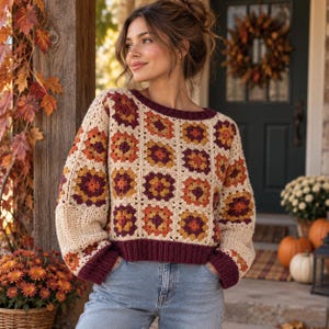 May include: A long-sleeved crochet sweater featuring a granny square pattern in shades of cream, orange, yellow, and dark maroon. The garment has a round neckline and ribbed cuffs and hem in a solid dark maroon yarn, paired with blue denim jeans.