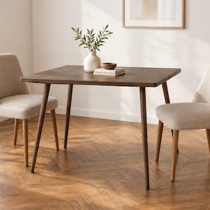Op de afbeelding: Een vierkante donkerbruine houten eettafel met vier taps toelopende poten, vergezeld door twee lichtbeige gestoffeerde stoelen met houten poten. Een witte keramische vaas met groen blad en een kleine houten kom staan op het tafeloppervlak.