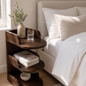 Puede incluir: Una mesa auxiliar de madera de color marrón oscuro con tres niveles y estantes curvos. El estante superior sostiene un jarrón de cerámica beige con follaje verde y un vaso transparente. El estante central contiene dos libros y un cuenco blanco.
