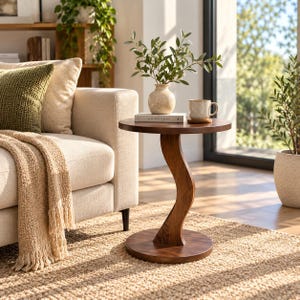 Puede incluir: Una mesa auxiliar redonda de madera con una base de pedestal curva en forma de S y una base circular plana. La mesa presenta un acabado de grano de madera marrón oscuro liso y se sostiene sobre una superficie plana, con un jarrón y una taza.