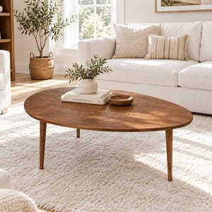 May include: An oval coffee table made of medium-brown wood with three tapered legs. On the surface sits a white textured vase containing green leaves, a stack of two books with the word The Kinfolk printed on the spine, and a small wooden bowl.