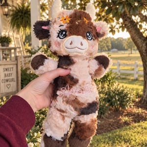 May include: A plush cow toy featuring a cream and brown spotted pattern, embroidered facial features with large blue eyes, small horns, and orange flower accents on the head. The chest area displays cursive pink embroidery of the name Amelia.