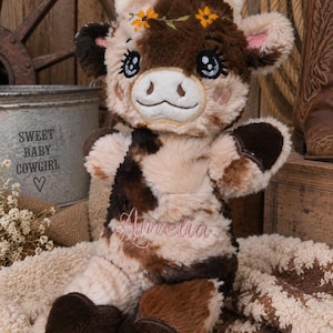 May include: A plush cow toy with brown and cream patches, featuring embroidered eyes, a floral crown on its head, and the name Amelia stitched in pink thread on its chest. The stuffed animal has dark brown hooves and soft, textured fur.