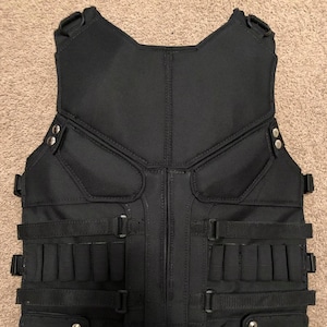 Può includere: Un gilet tattico nero caratterizzato da un pannello frontale strutturato con sezioni imbottite, molteplici cinghie orizzontali e passanti integrati. Il capo include due tasche rettangolari inferiori fissate con bottoni a pressione in metallo argentato.