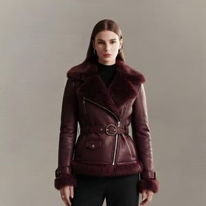 May include: A deep burgundy leather jacket featuring a wide faux fur collar, matching fur cuffs, and a belted waist with a circular buckle. The jacket has an asymmetrical silver zip closure, a small flap pocket, and is paired with black trousers.