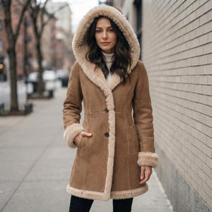 Può includere: Un cappotto in camoscio color marrone chiaro con un ampio cappuccio foderato in pelliccia sintetica color crema. Il cappotto presenta una chiusura frontale con bottoni, maniche lunghe con polsini in pelliccia e un orlo rifinito in pelliccia.