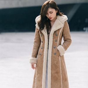 May include: A long, tan-colored coat featuring a thick, cream-colored faux shearling lining along the front opening, hood, and cuffs. The coat has a double-breasted button closure with circular buttons and a structured, full-length silhouette.