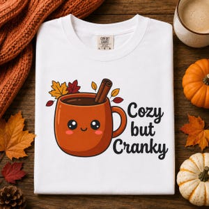 May include: A white t-shirt featuring a graphic of a cute, smiling orange mug filled with a dark beverage and a cinnamon stick. The mug is surrounded by colorful autumn leaves. The text "Cozy but Cranky" is printed in black next to the mug.