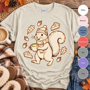 Peut inclure: Un t-shirt couleur sable avec une illustration graphique d'un écureuil portant un bonnet et une écharpe, tenant une tasse de café. Le motif comprend des éléments environnants de glands, de feuilles de chêne et deux parts de tarte.