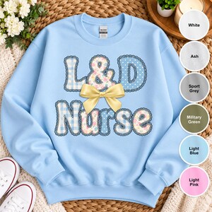 May include: A light blue crewneck sweatshirt featuring the text L&D Nurse in a scalloped-edge font. The letters have a mix of blue gingham, white polka dot, and floral patterns. A large, solid yellow bow is centred over the text.