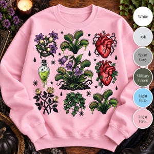 May include: A light pink crewneck sweatshirt featuring a graphic print of botanical and anatomical illustrations, including Venus flytraps, purple bell flowers, yellow roses, red human hearts, a green potion bottle, and small black spiders.