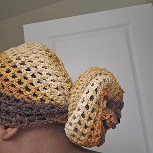 May include: A crocheted head covering in shades of brown, tan, and gold. The open weave design allows for breathability. The head covering is styled in a bun shape.