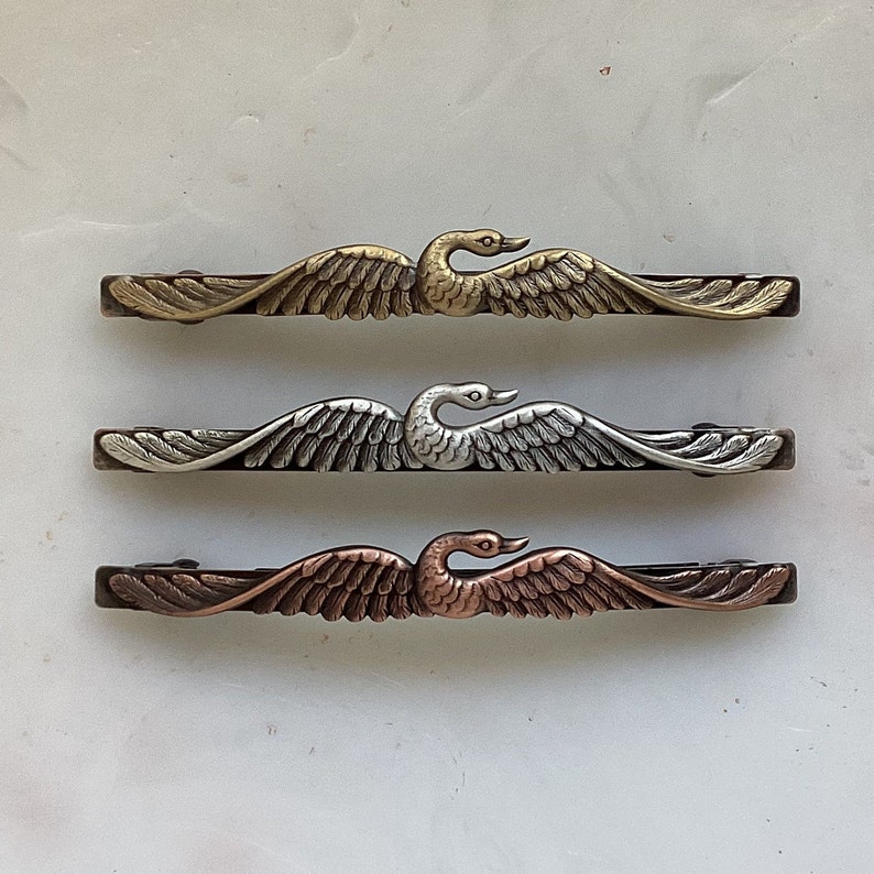 K&ouml;nnte beinhalten: Drei dekorative Haarklammern aus Metall in Form eines Schwans in Gold-, Silber- und Kupferausf&uuml;hrung. Jede Klammer zeigt ein detailliertes Schwanendesign mit ausgebreiteten Fl&uuml;geln.