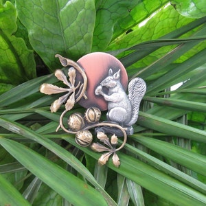 May include: A detailed metal squirrel brooch with a silver body, holding a nut, set against a copper-colored circle. The brooch features bronze-colored leaves and acorns, all resting on a bed of green leaves.