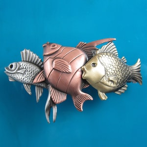 Puede incluir: Tres figuritas de peces de metal en fila. El pez de la izquierda es plateado, el del medio es de cobre y el de la derecha es dorado. Los peces est&aacute;n todos mirando hacia el lado derecho de la imagen.