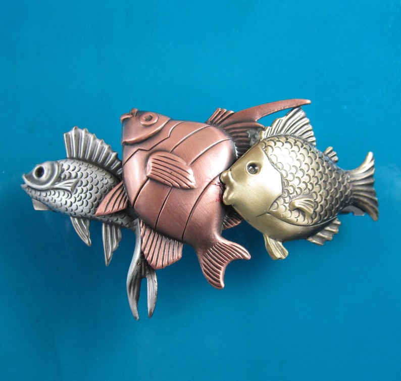 Puede incluir: Tres figuritas de peces de metal en fila. El pez de la izquierda es plateado, el del medio es de cobre y el de la derecha es dorado. Los peces est&aacute;n todos mirando hacia el lado derecho de la imagen.