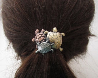 May include: A hair tie featuring three turtle charms. The turtles are made of metal and are in shades of gold, silver, and copper.