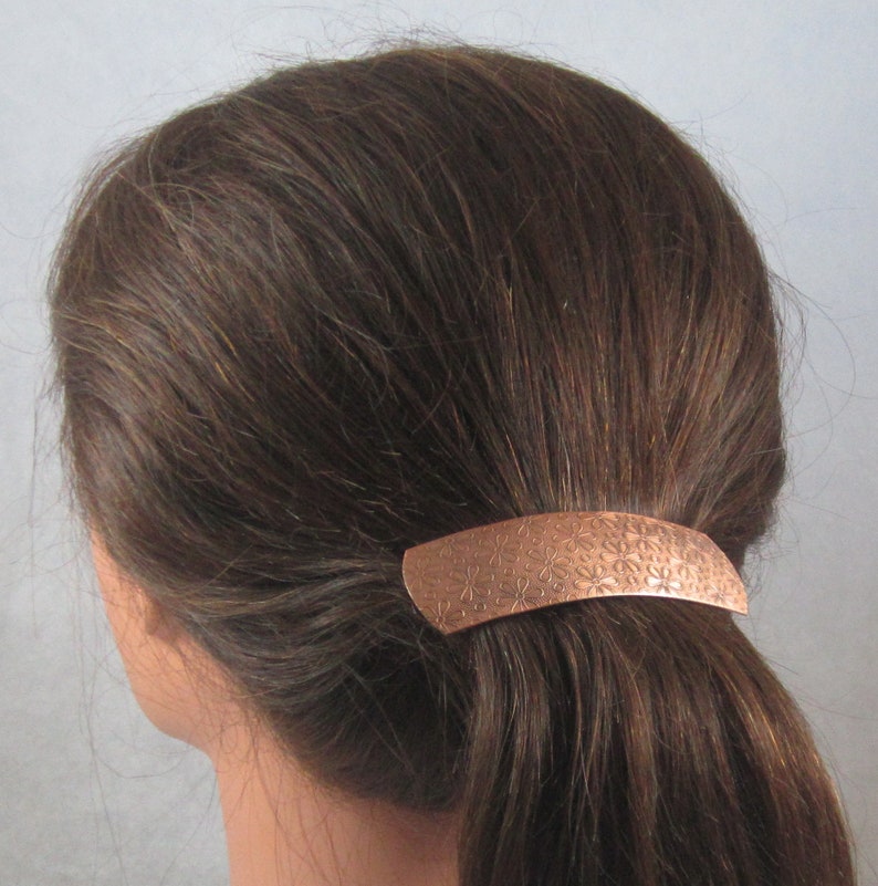 Puede incluir: Una pinza para el pelo de cobre con un dise&ntilde;o floral. La pinza mide aproximadamente 5 cm de largo y se muestra puesta en una coleta.