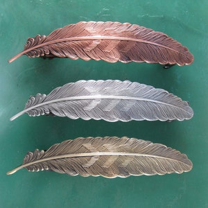 May include: Three feather-shaped hair clips in different finishes: copper, silver, and gold. Each clip has a detailed feather design.