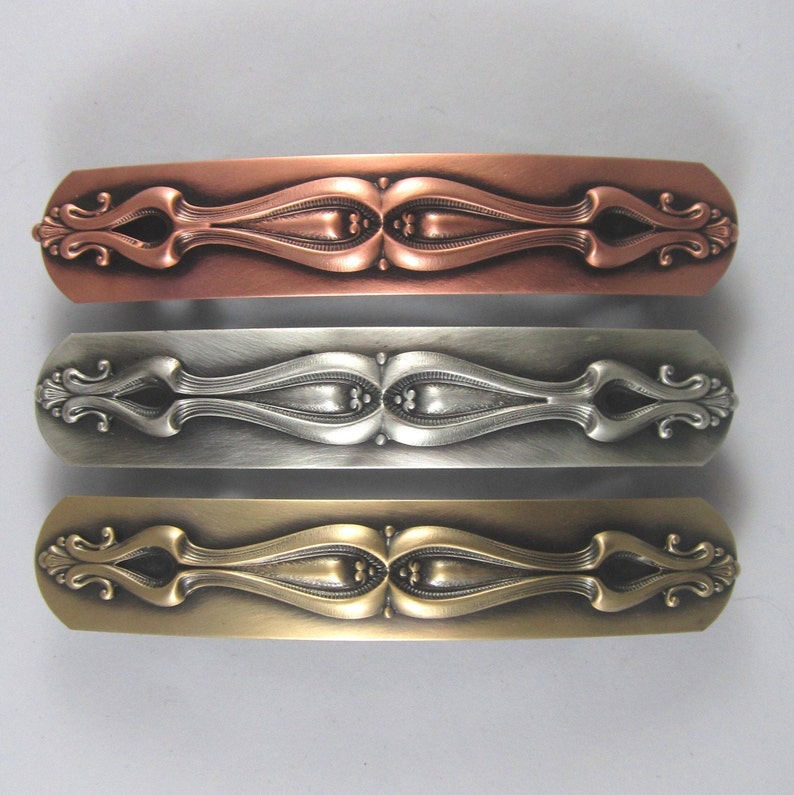 K&ouml;nnte beinhalten: Drei verzierte Haarklammern in verschiedenen Ausf&uuml;hrungen: Kupfer, Silber und Gold. Jede Klammer hat ein dekoratives, symmetrisches Design mit einer zentralen ovalen Form und filigranen Schn&ouml;rkeln.