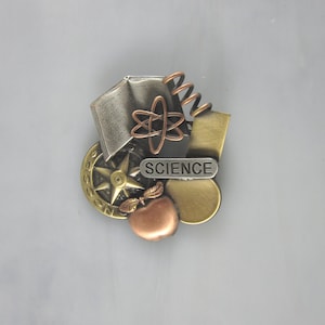 May include: A metal pin with a book, compass, apple, and atom design. The pin says "SCIENCE" on it.
