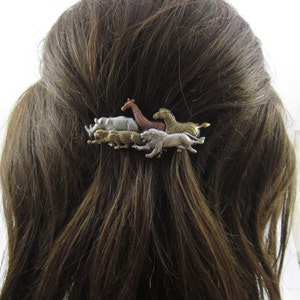 May include: A silver, gold, and bronze hair clip featuring a line of five safari animals: a rhinoceros, a lion, a zebra, a giraffe, and a cheetah. The animals are all facing the same direction and are arranged in a line.