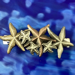 May include: A cluster of five gold and bronze starfish hair clips. The starfish are arranged in a staggered pattern, with the largest starfish at the bottom and the smallest starfish at the top.