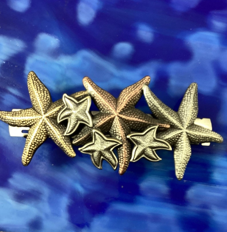 May include: A cluster of five gold and bronze starfish hair clips. The starfish are arranged in a staggered pattern, with the largest starfish at the bottom and the smallest starfish at the top.