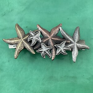May include: A set of five metal starfish hair clips in gold, silver, and bronze tones. The starfish are arranged in a cluster, with the largest starfish at the bottom and the smallest starfish at the top.