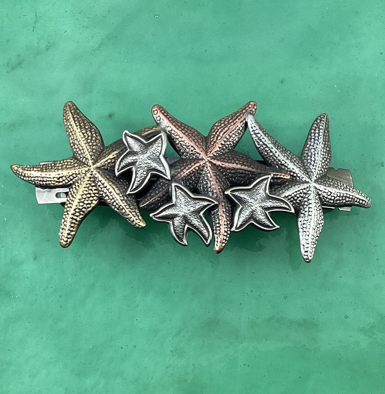 May include: A set of five metal starfish hair clips in gold, silver, and bronze tones. The starfish are arranged in a cluster, with the largest starfish at the bottom and the smallest starfish at the top.