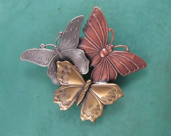 May include: Three metal butterfly brooches in silver, gold, and copper tones. The butterflies are arranged in a cluster, with the silver butterfly on top and the copper butterfly on the bottom.