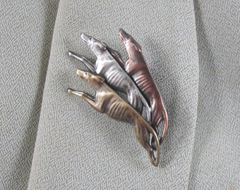 Pode incluir: Broche com três figuras estilizadas de galgos em tons de bronze, prata e cobre. Os cães estão saltando, com os corpos arqueados e as cabeças levantadas. O broche está preso a um tecido verde claro texturizado.