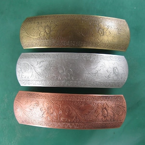 Könnte beinhalten: Drei Haarspangen aus Metall in Gold, Silber und Kupfer. Jede Spange hat eine gebogene Form und ist mit einem geprägten Blumen- und Rankenmuster verziert. Die Spangen werden auf einer grünen Oberfläche präsentiert.