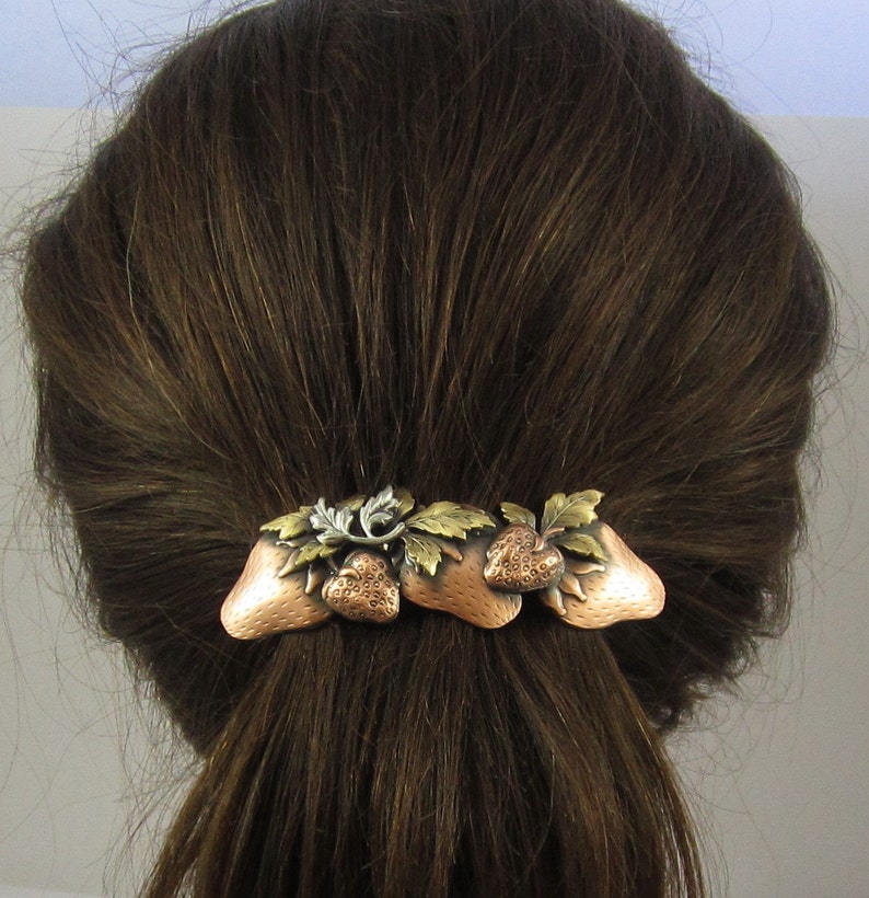Puede incluir: Una pinza para el pelo de cobre y plata con un dise&ntilde;o de tres fresas y hojas.